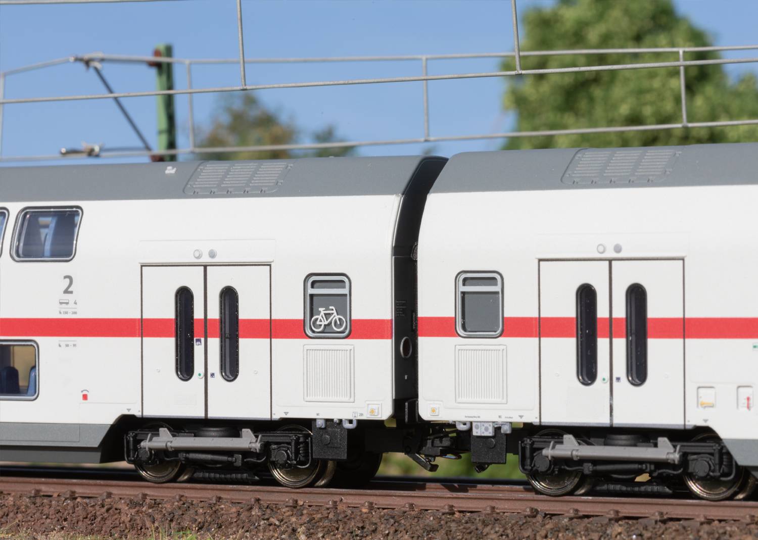 A white train with a red stripe on railway tracks. The bicycle symbol indicates an area for bicycles. Trees are visible in the background.