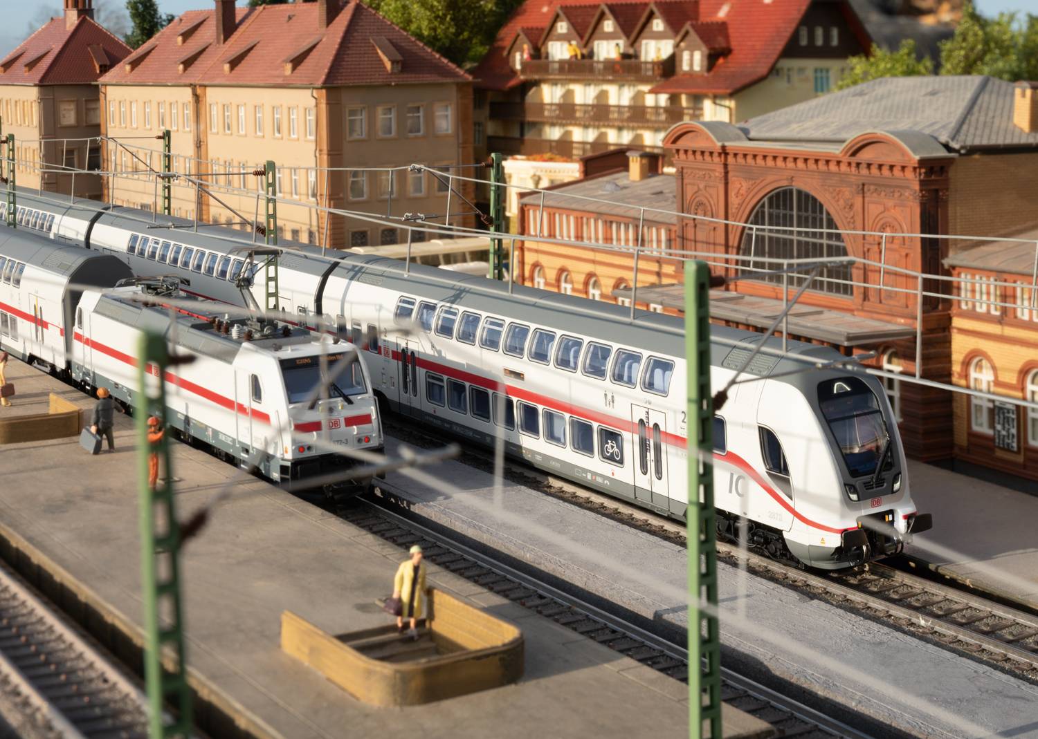 A model railway landscape shows a station with two trains at the tracks. A passenger is standing on the platform.