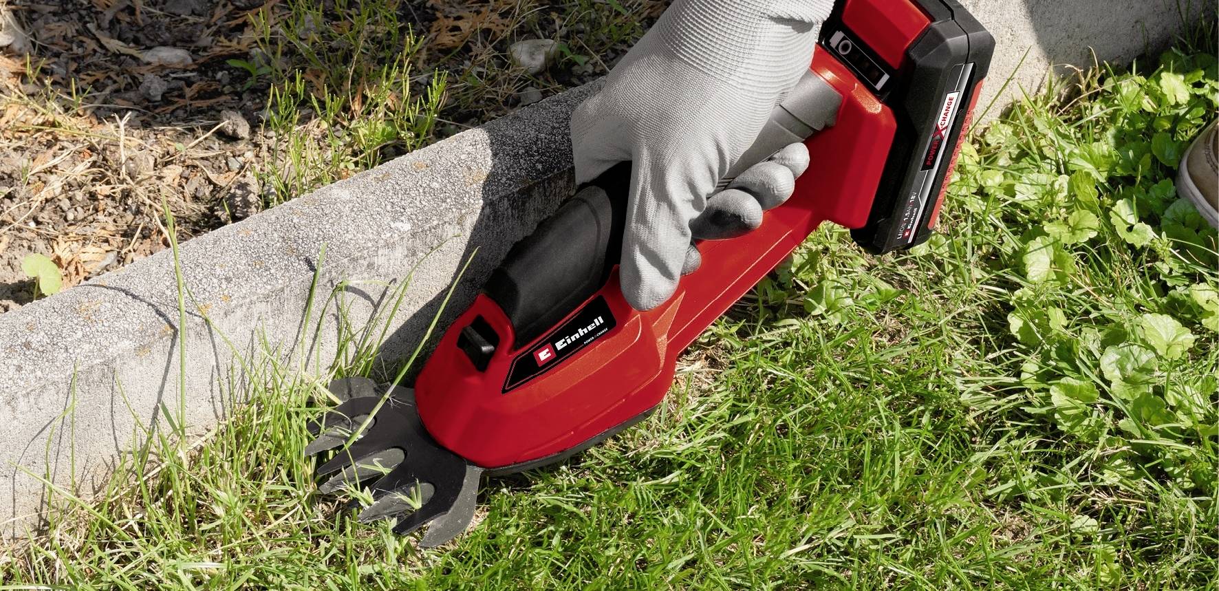 A person is cutting grass along a kerb edge using a red cordless grass trimmer.