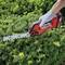 A person is holding a red hedge trimmer and cutting a lush green hedge. The person is wearing grey gloves.