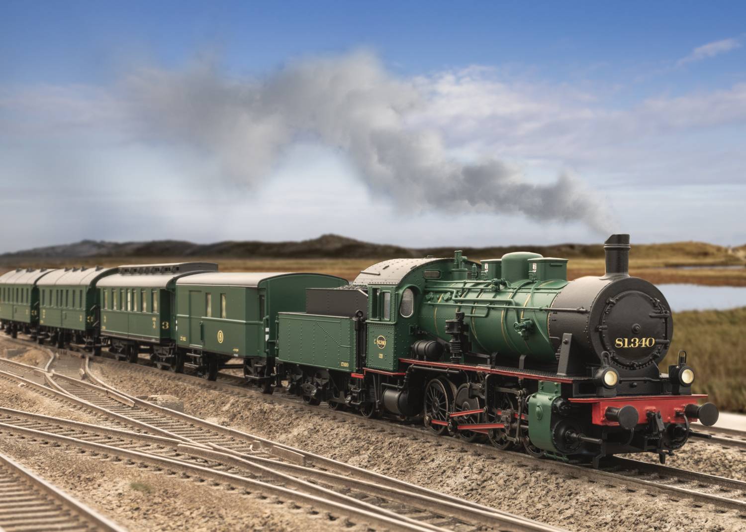 Green steam locomotive with several carriages travels along railway tracks through rural countryside under a blue sky with clouds.