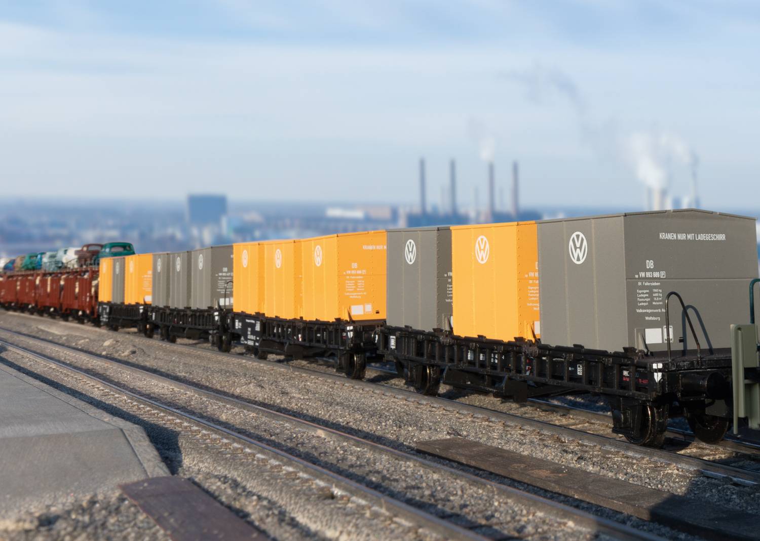 Several goods wagons with orange-grey containers bearing the VW brand are stationed on a railway track, with chimneys visible in the background.