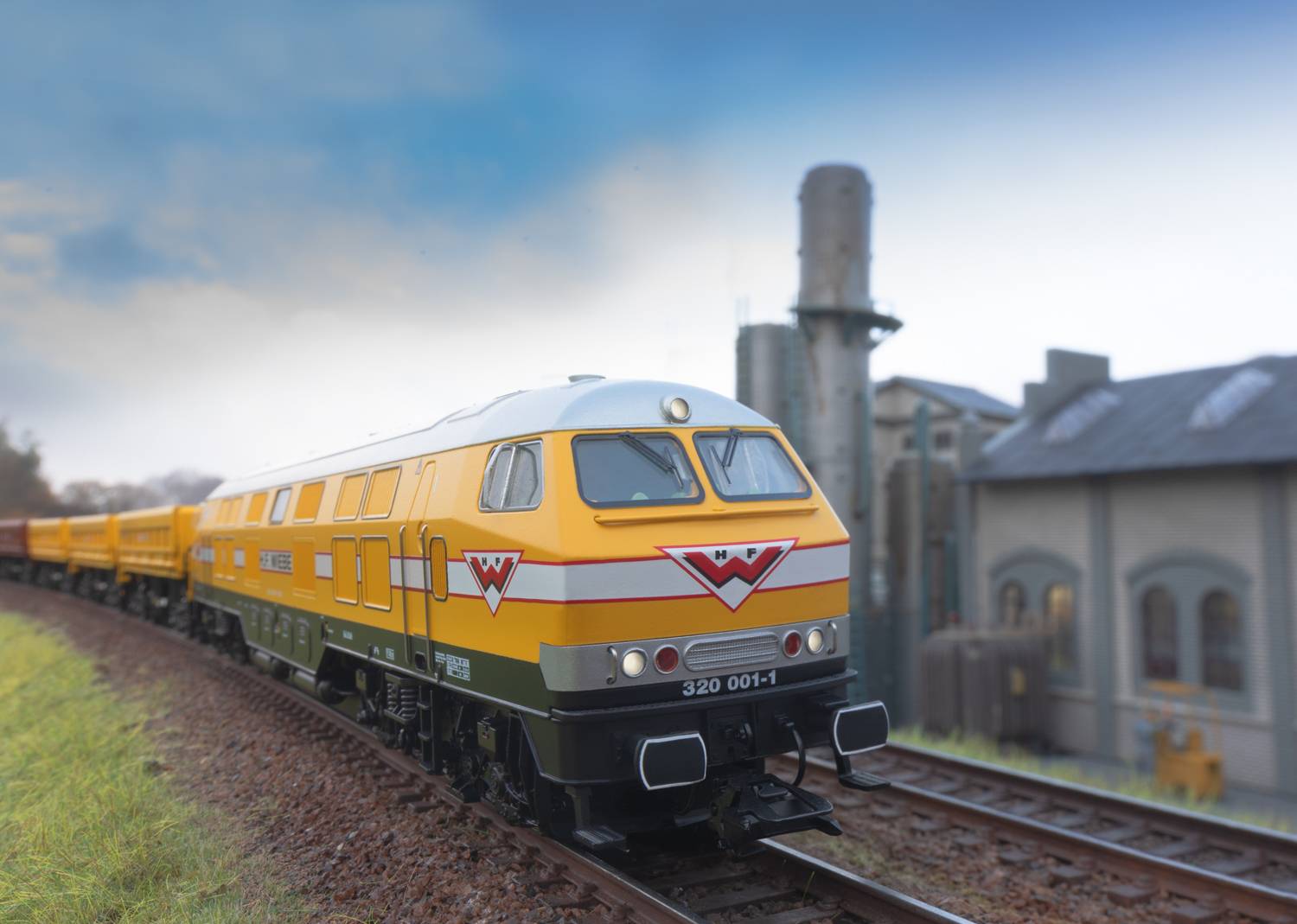 A yellow train on a railway track, with an industrial building in the background. The sky is cloudy, and the surroundings are green.