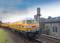 A yellow train on a railway track, with an industrial building in the background. The sky is cloudy, and the surroundings are green.