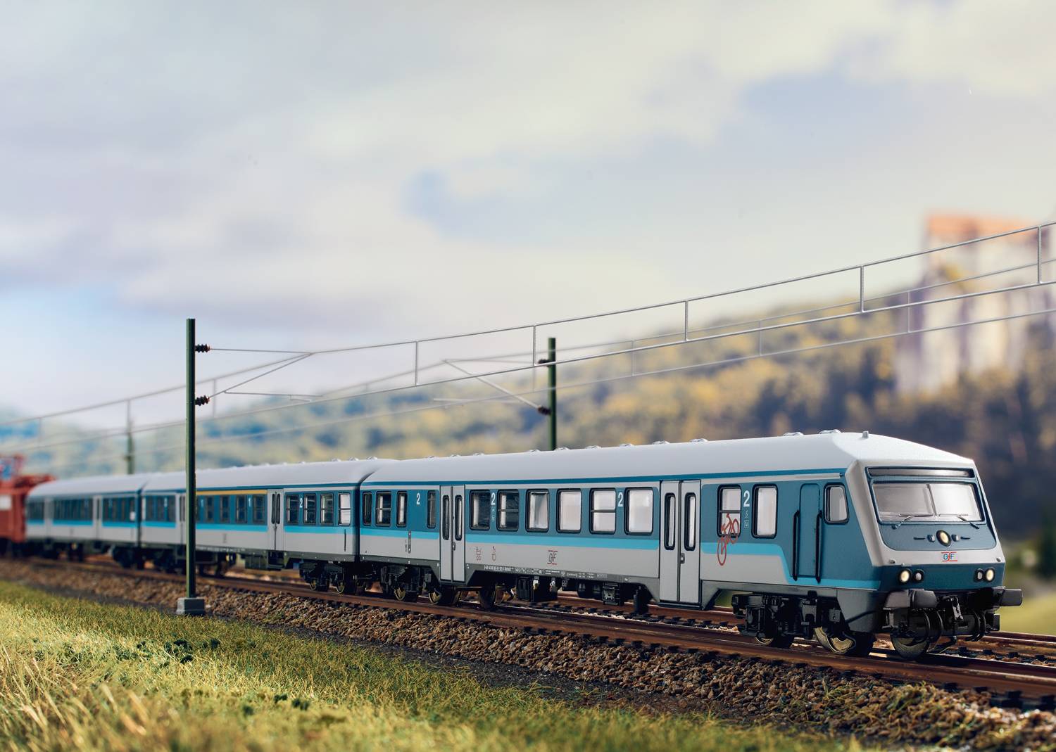 A blue train travels along tracks on a route through a rural landscape. Electricity lines run parallel to the railway line.