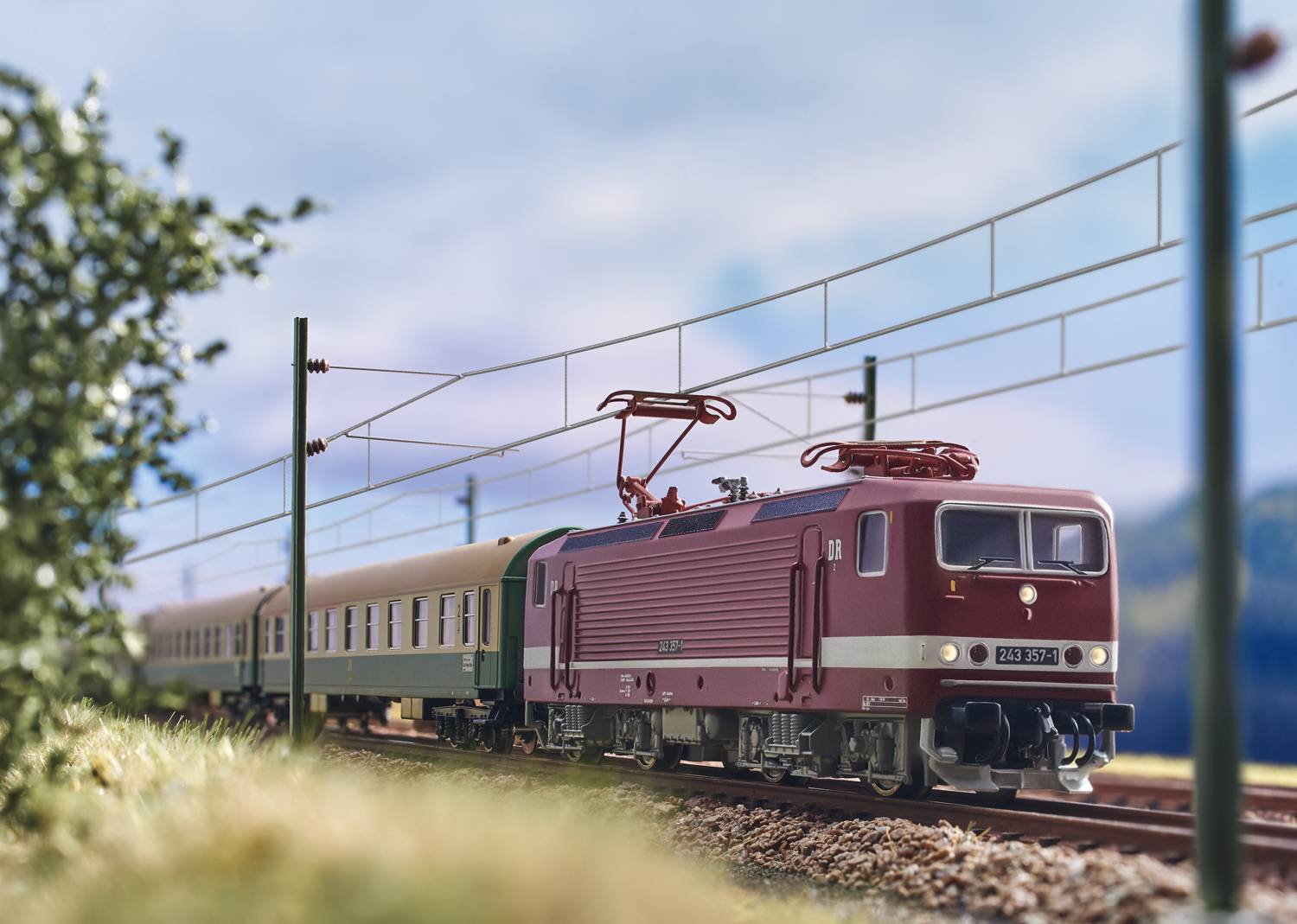 A red locomotive is pulling a green carriage along an electrified railway line through a rural, verdant landscape.