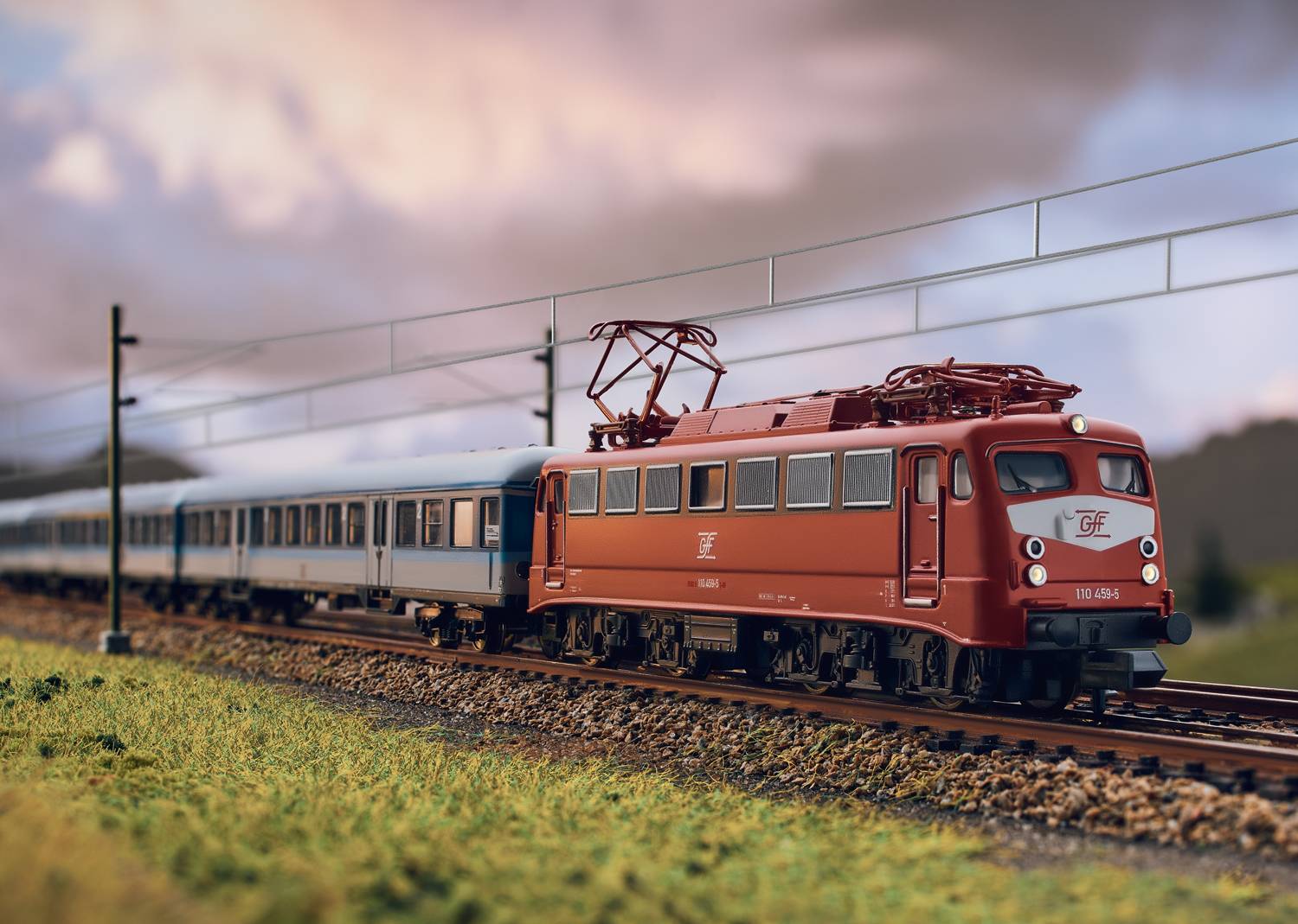 A red locomotive pulls several blue carriages along a model railway track, surrounded by green grass and a cloudy sky in the background.