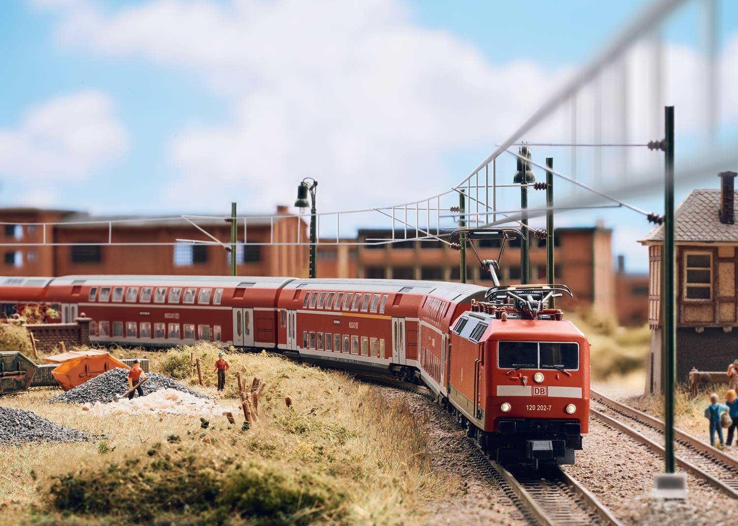 Model railway scene with a red train travelling along a track through a miniature landscape. In the background are buildings and workers.
