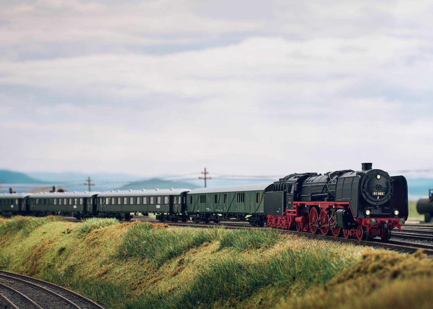 A realistic model of a steam locomotive pulls several carriages across a green, hilly landscape beneath a cloudy sky.