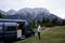 A person is standing next to an open camper van, looking out at wooded hills and tall, rocky mountains in the background.