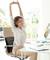A woman in an elegant office outfit stretches at her desk in front of a computer screen. A bright, modern office space is visible in the background.
