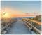 Wooden walkway leads to the beach, framed by sand dunes, at sunset. Birds fly across the sky, calm sea in the background.