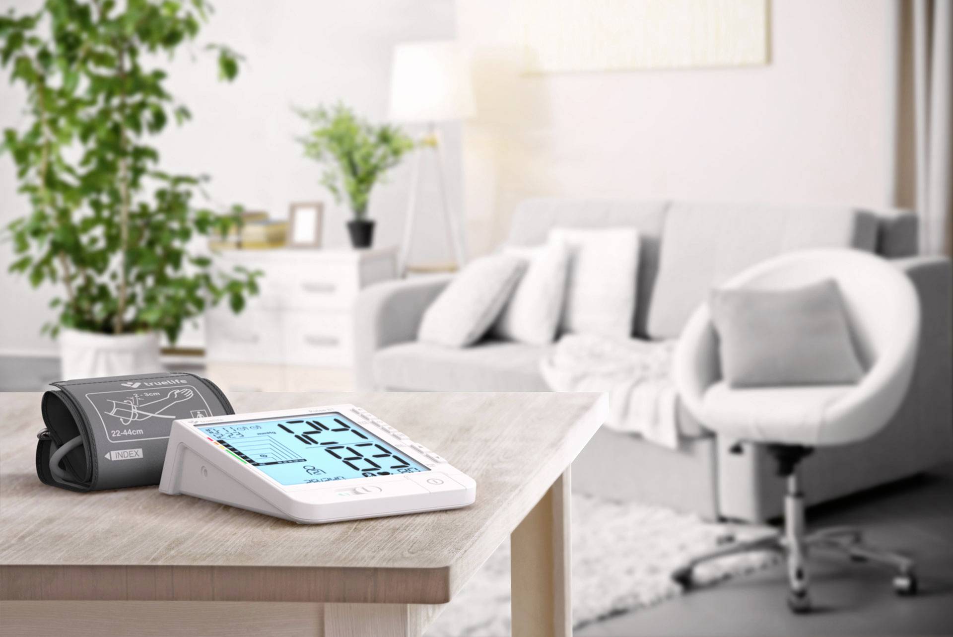 Blood pressure monitor on a table in a modern living room, an armchair and a large potted plant to the left, with a lamp in the background.