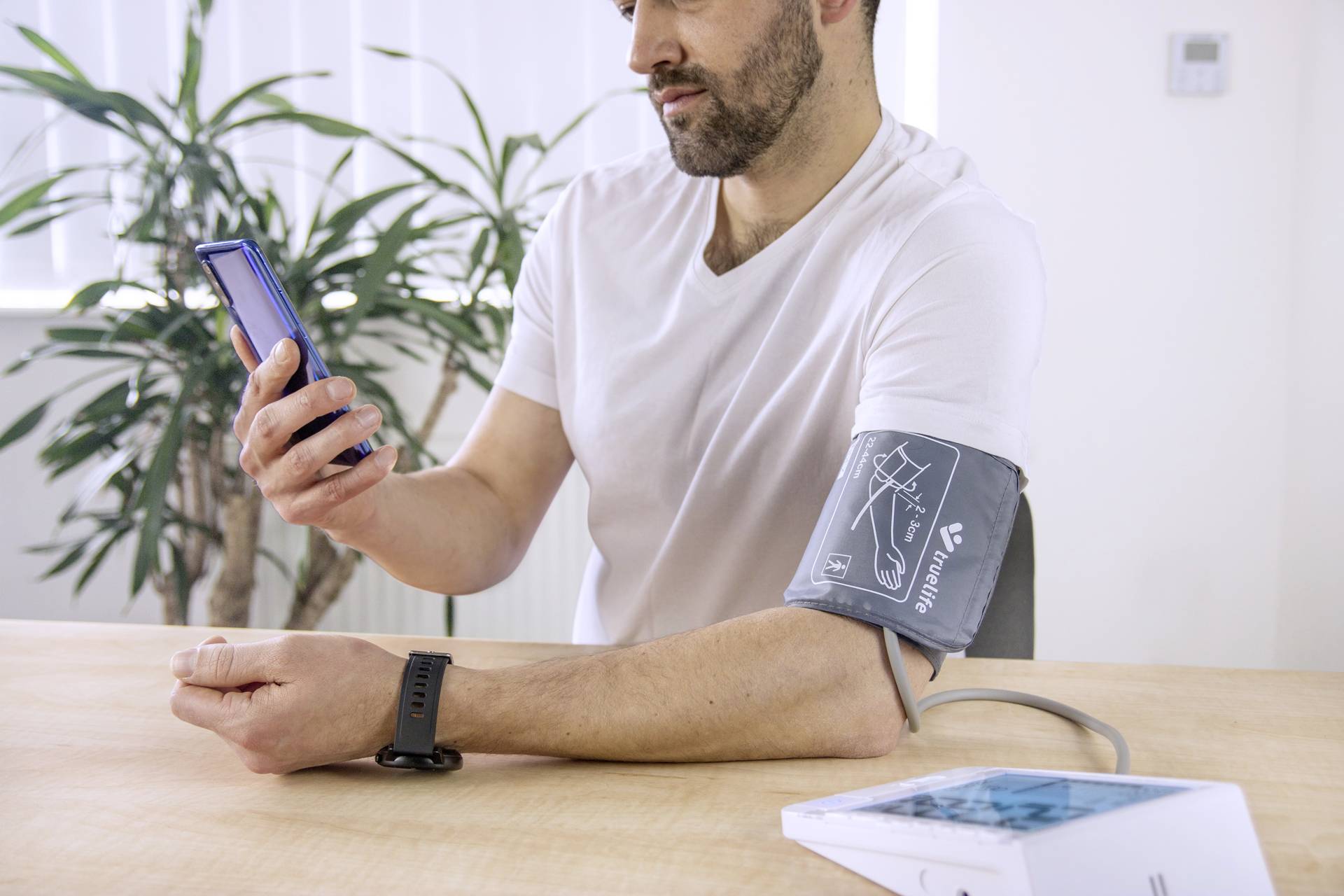 A person is measuring their blood pressure with a digital device. They are looking at their mobile phone. A plant is visible in the background.