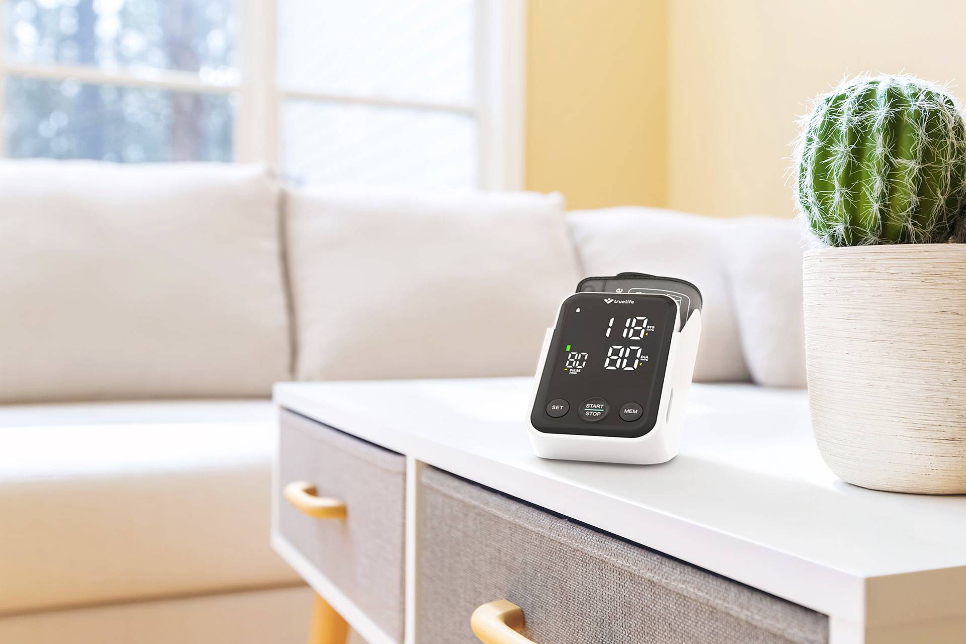 A digital blood pressure monitor on a white table in front of a sofa, next to a small cactus in a pot.