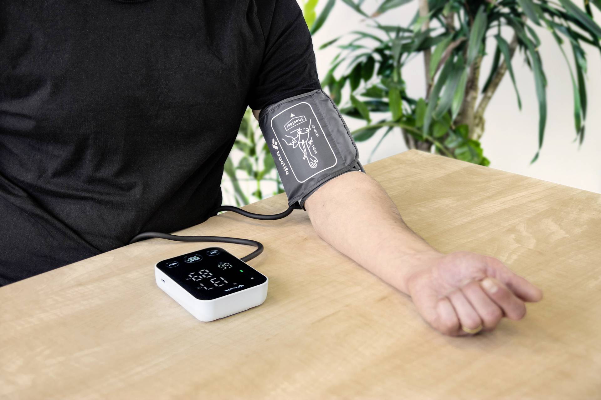 A person is measuring their blood pressure using a digital blood pressure monitor on their arm. Green plants can be seen in the background.