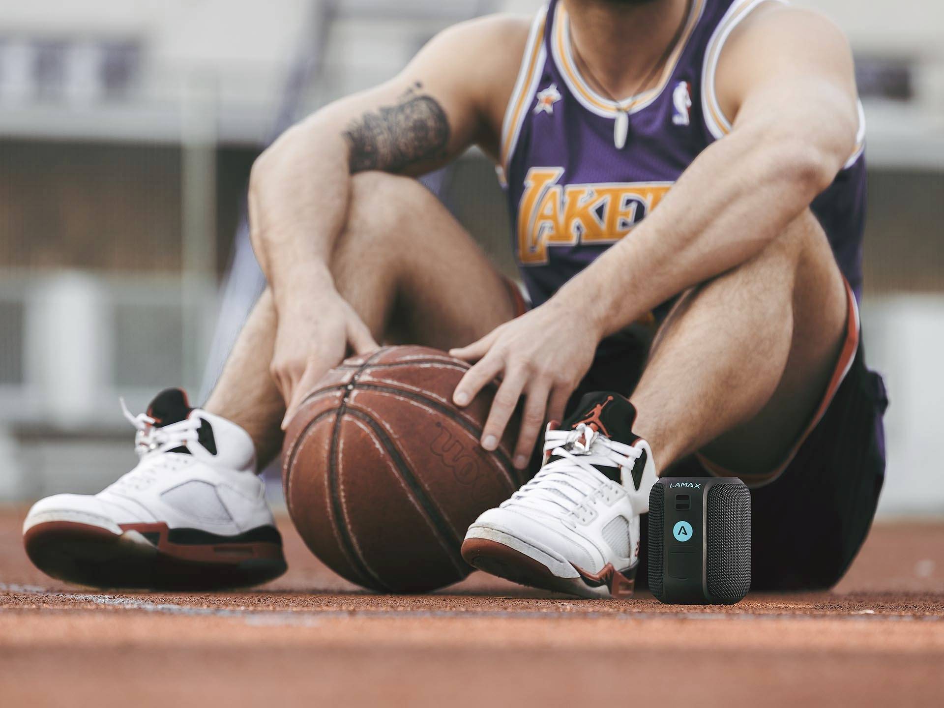 A man in sportswear is sitting on a basketball court, holding a basketball. A small, black device is beside him.