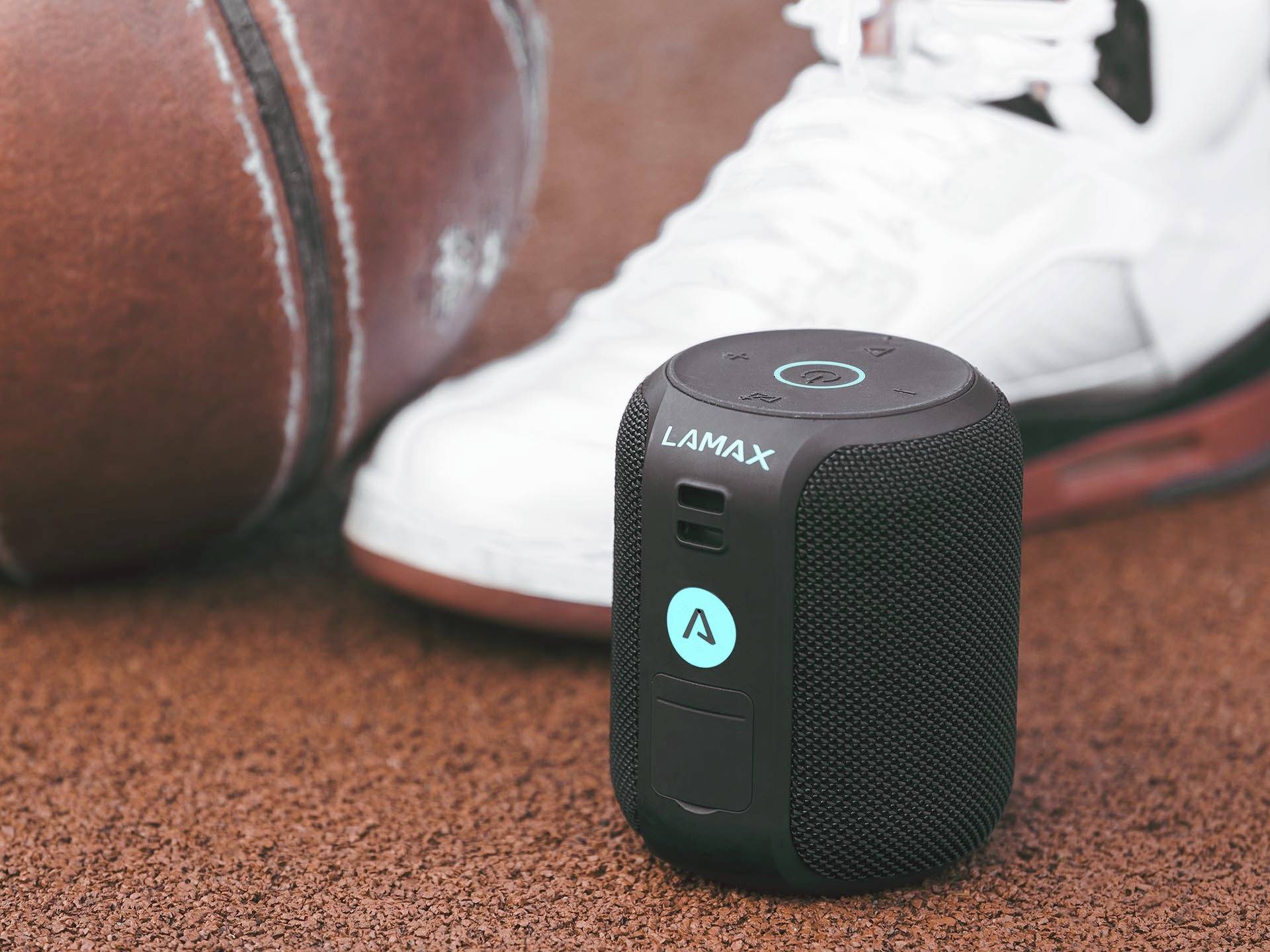 A portable speaker by Lamax is placed on a sports field floor next to a basketball and a trainers.