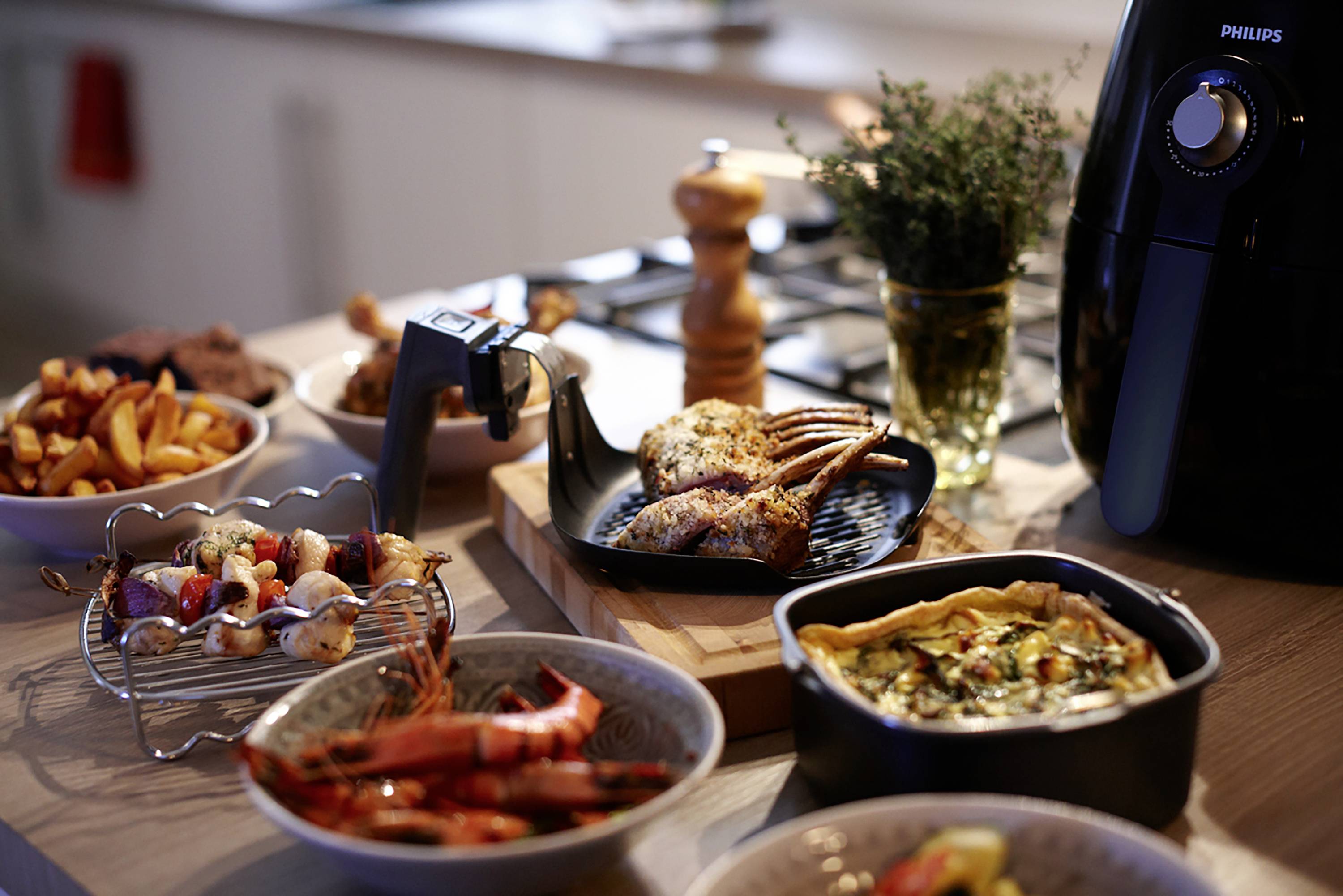A table is laid with various dishes: lamb chops on a griddle, prawns, vegetable quiche and chips. In the background stands an air fryer.