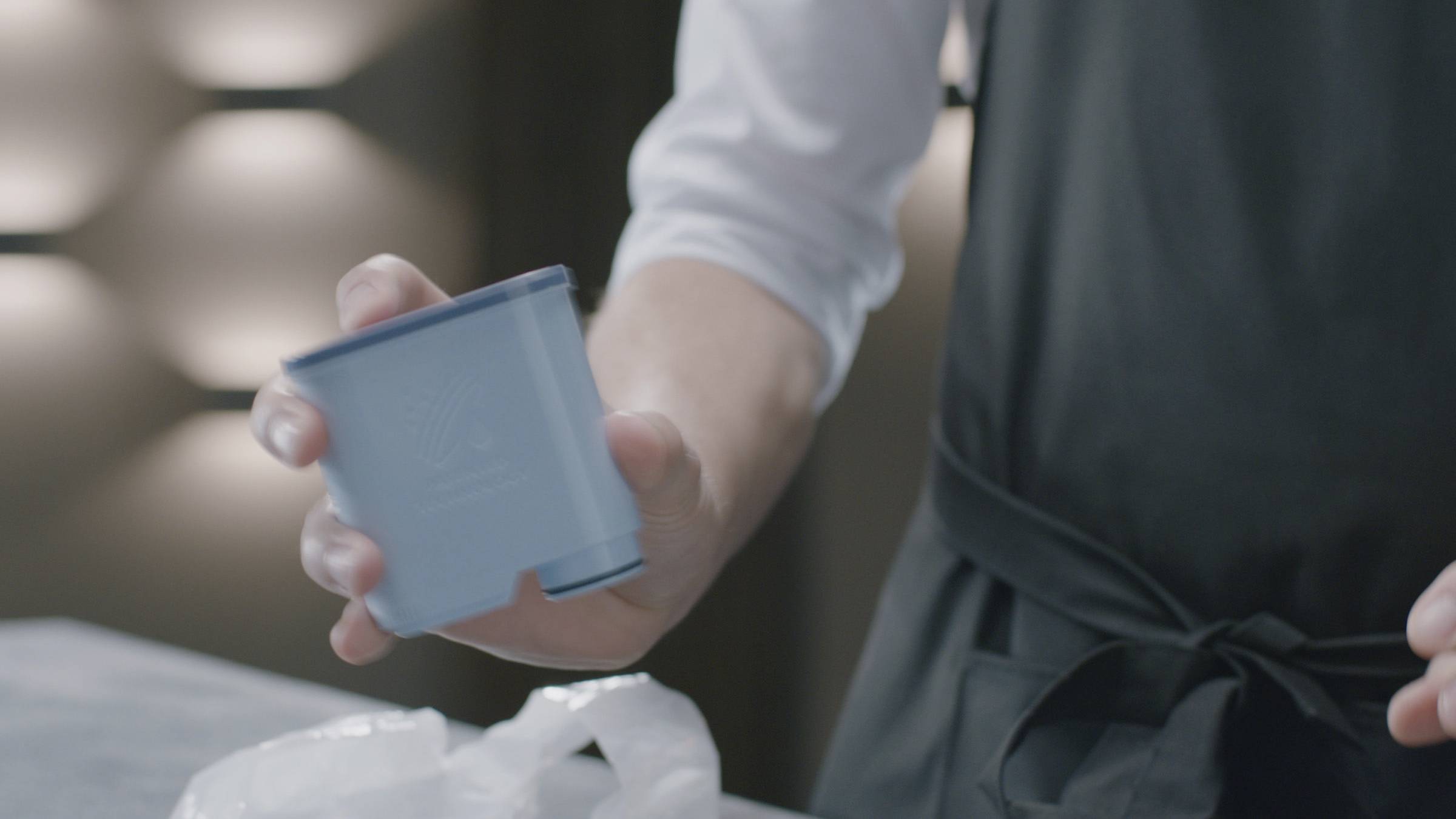 A person is holding a blue container in their hand. They are wearing an apron, and lights can be seen on a wall in the background.