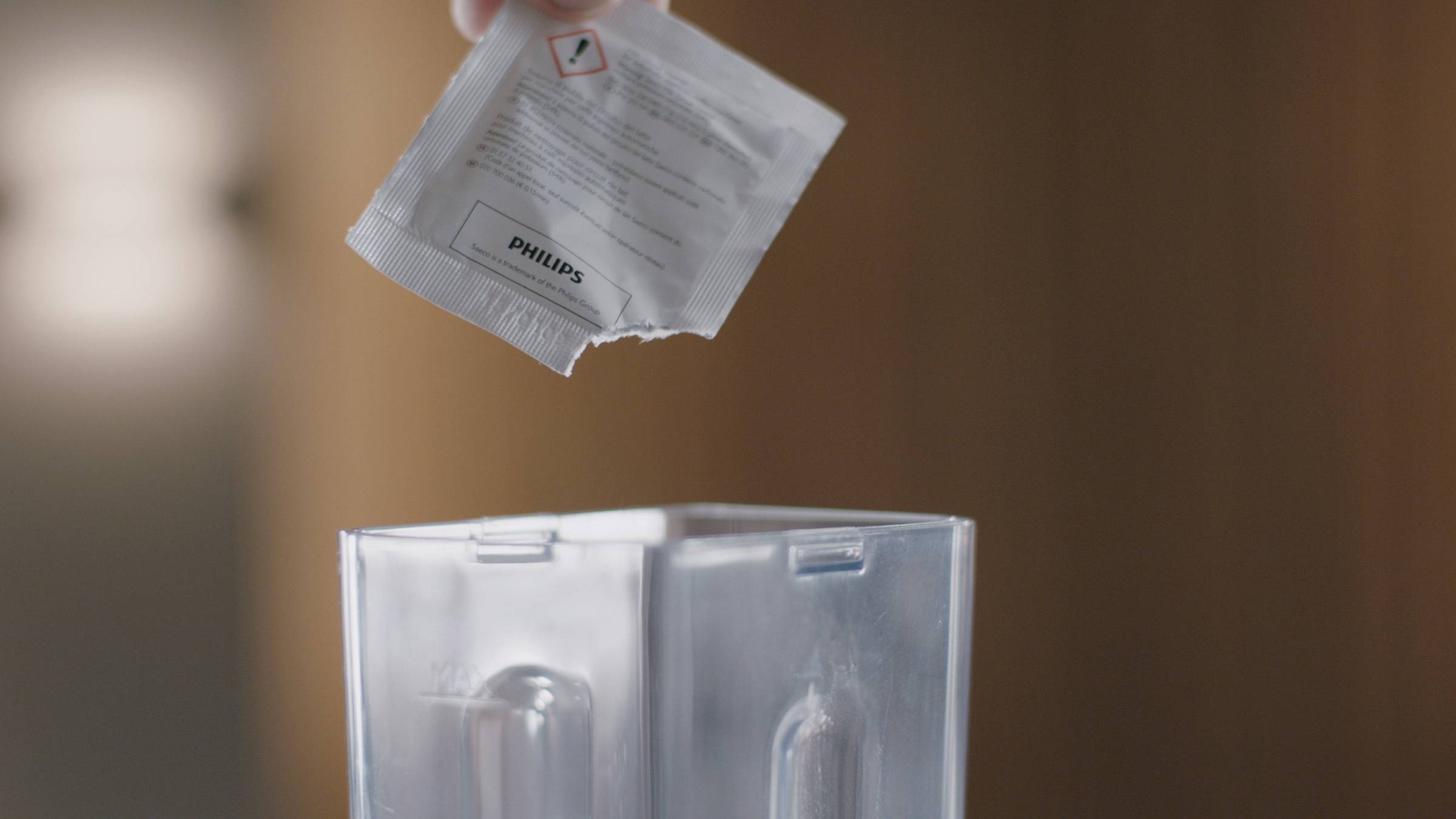 A hand is holding an open descaling powder packet over the water reservoir of a coffee machine, ready to pour.