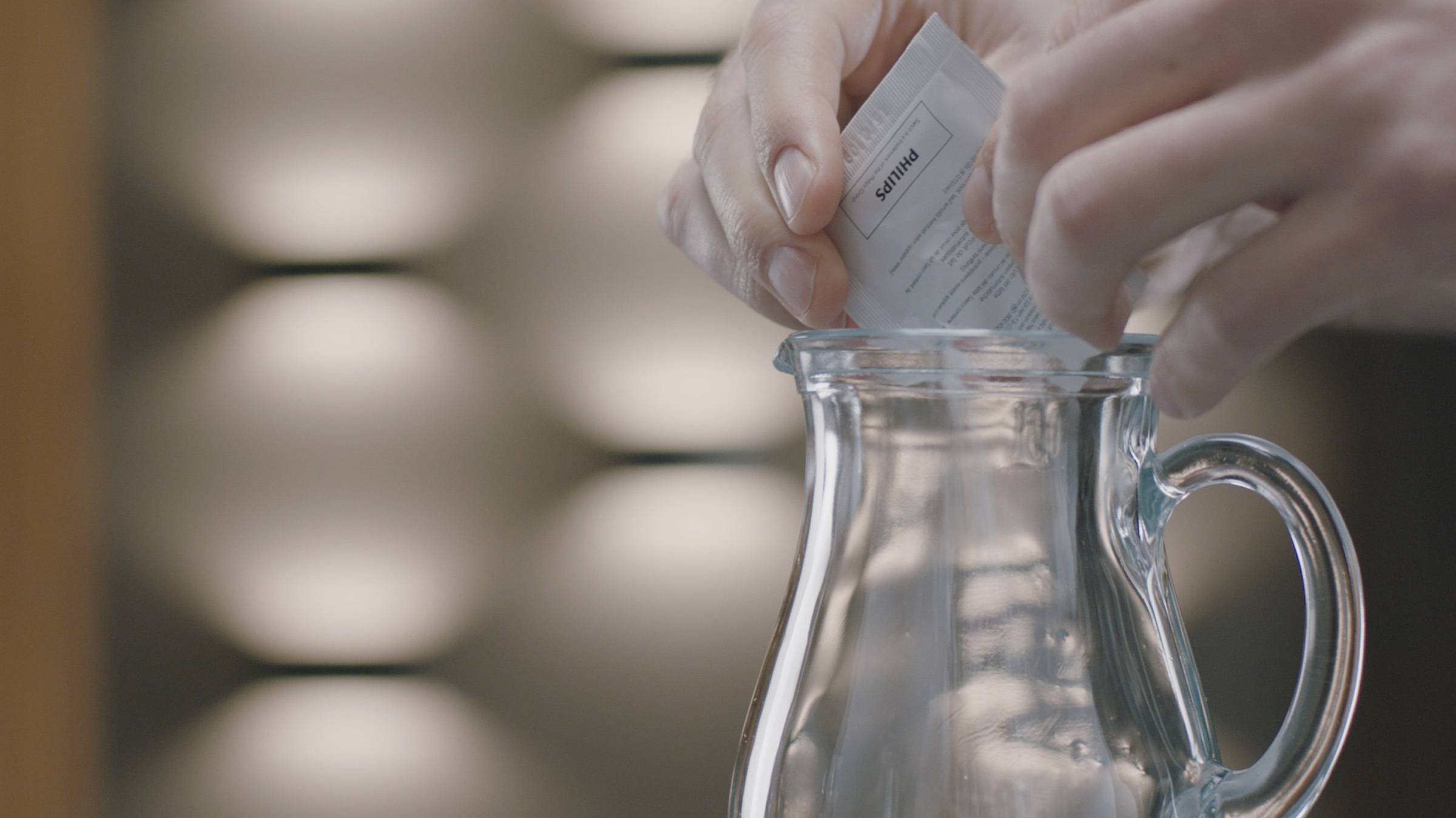 A person is pouring powder from a small sachet into an empty glass jar. Background slightly blurred.