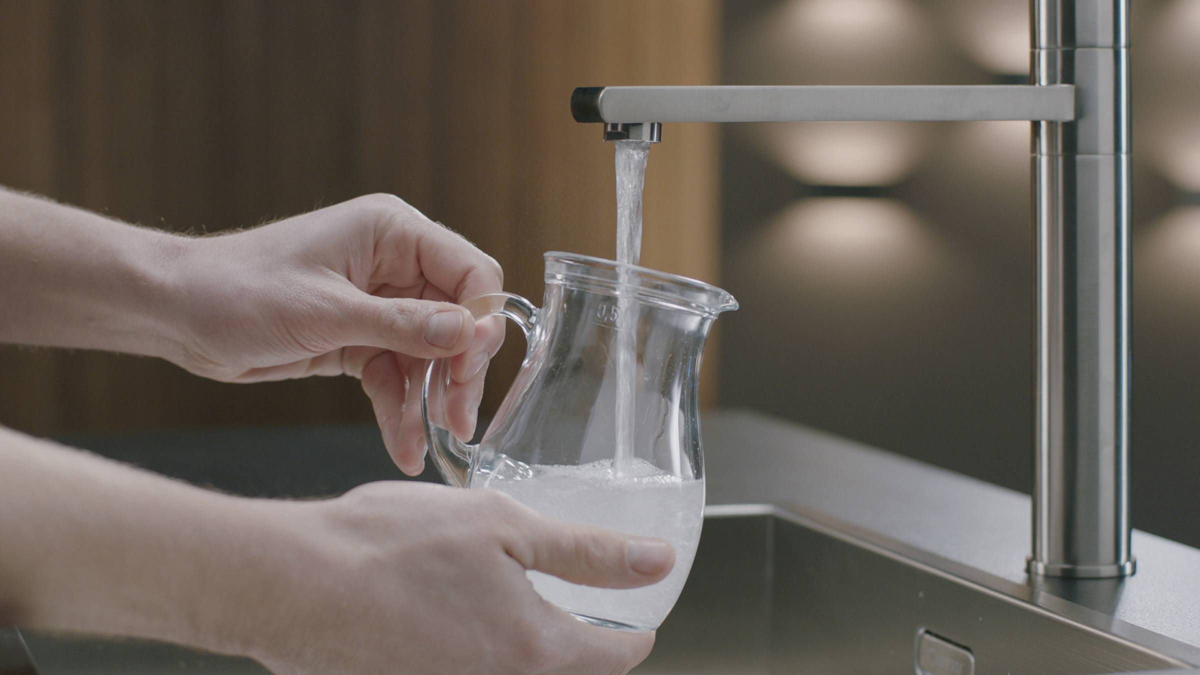 A person is holding a glass jug under a tap while it is being filled with clear water.