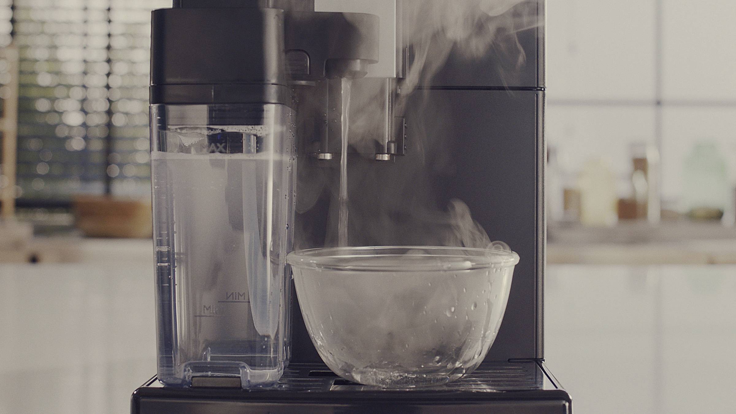 A black machine emits hot steam into a glass bowl. Blurred kitchen utensils can be seen in the background.