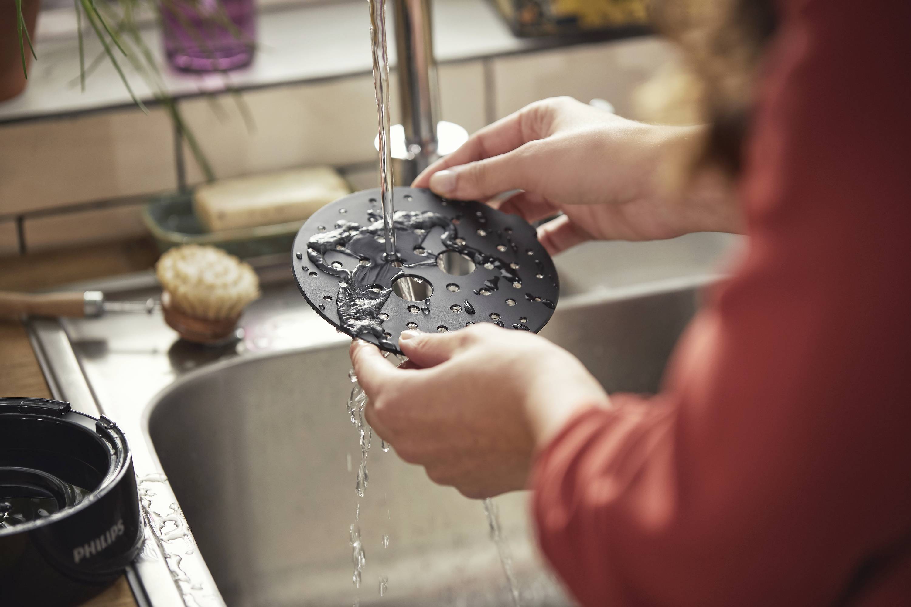 A person is rinsing a black metal disc under running water in the kitchen sink. Kitchen utensils can be seen in the background.