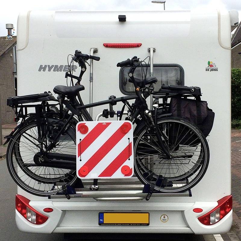 A bicycle carrier with two bicycles attached to the rear of a motorhome, parked on a road. A red and white warning triangle is visible.