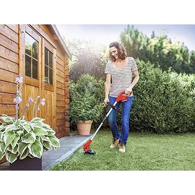 A woman is using a lawn trimmer to cut grass in a garden next to a wooden shed. Bushes are visible in the background.