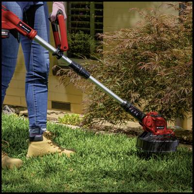 A person wearing jeans and work boots is using a red lawn trimmer to cut tall grass in the garden.