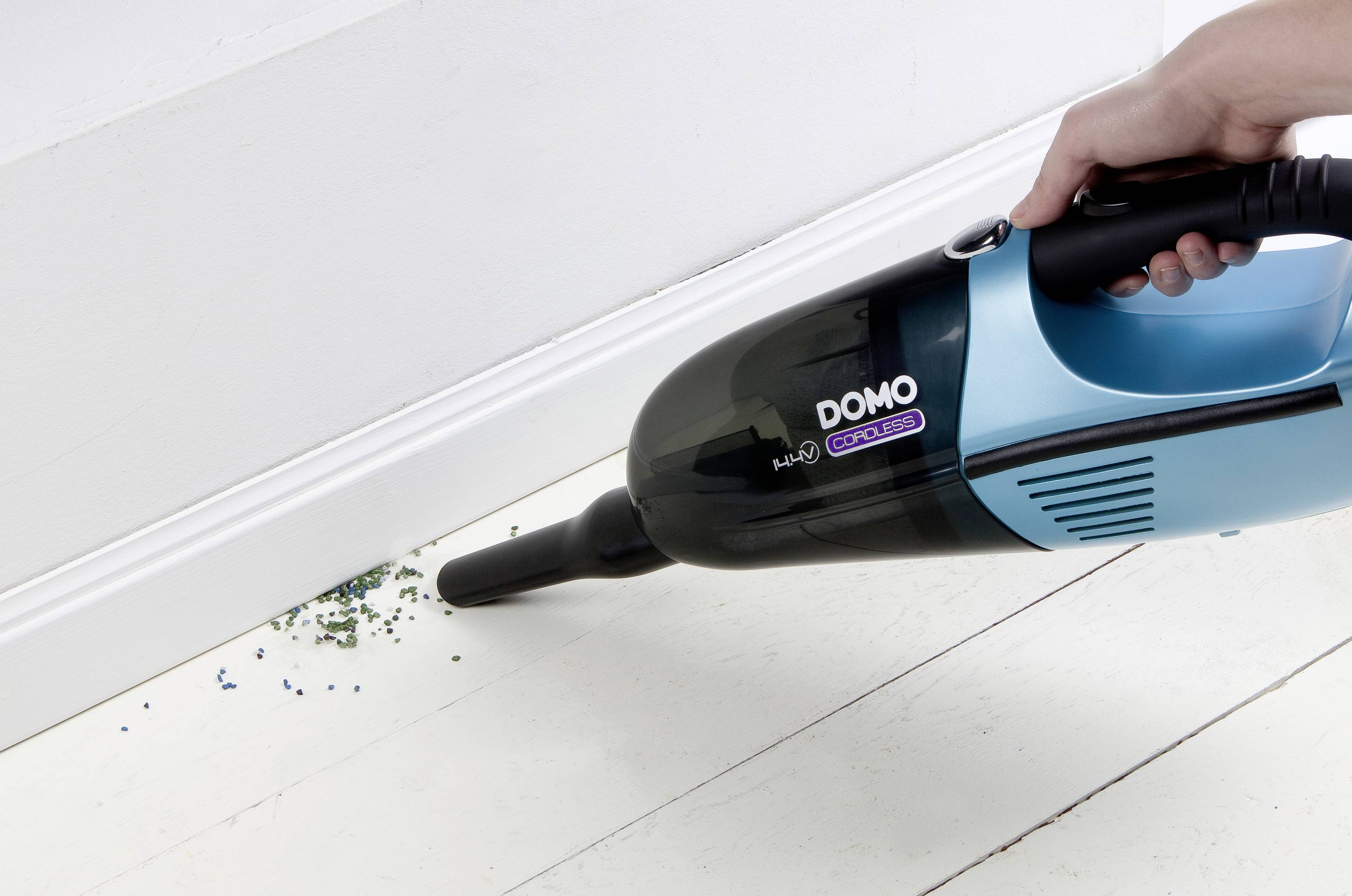 A hand is holding a blue handheld vacuum cleaner, which is sucking up blue and green crumbs from a white floor near the edge of a wall.