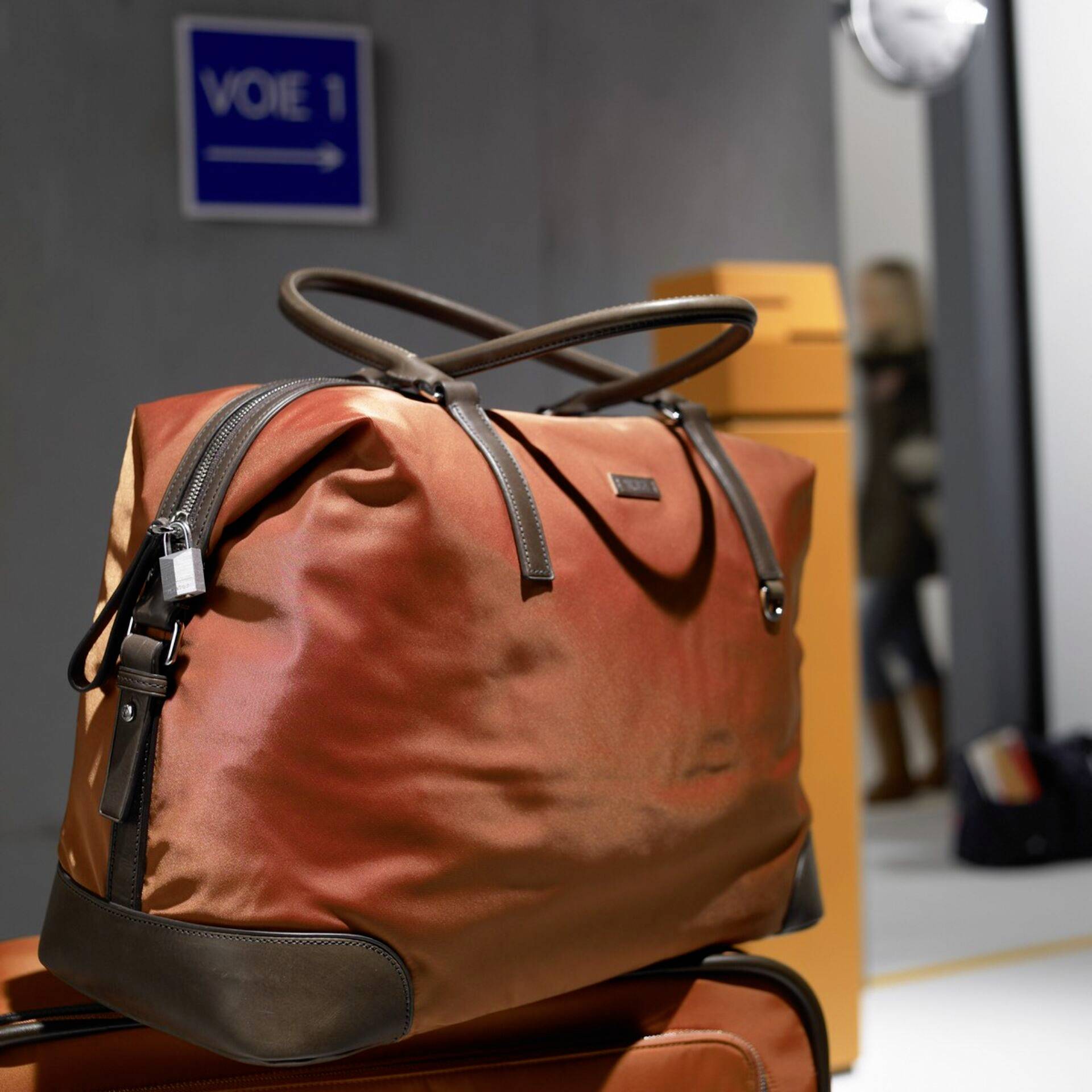Travel bag in warm colours on a suitcase, with a blurry figure and sign reading 'VOIE 1' in the background.