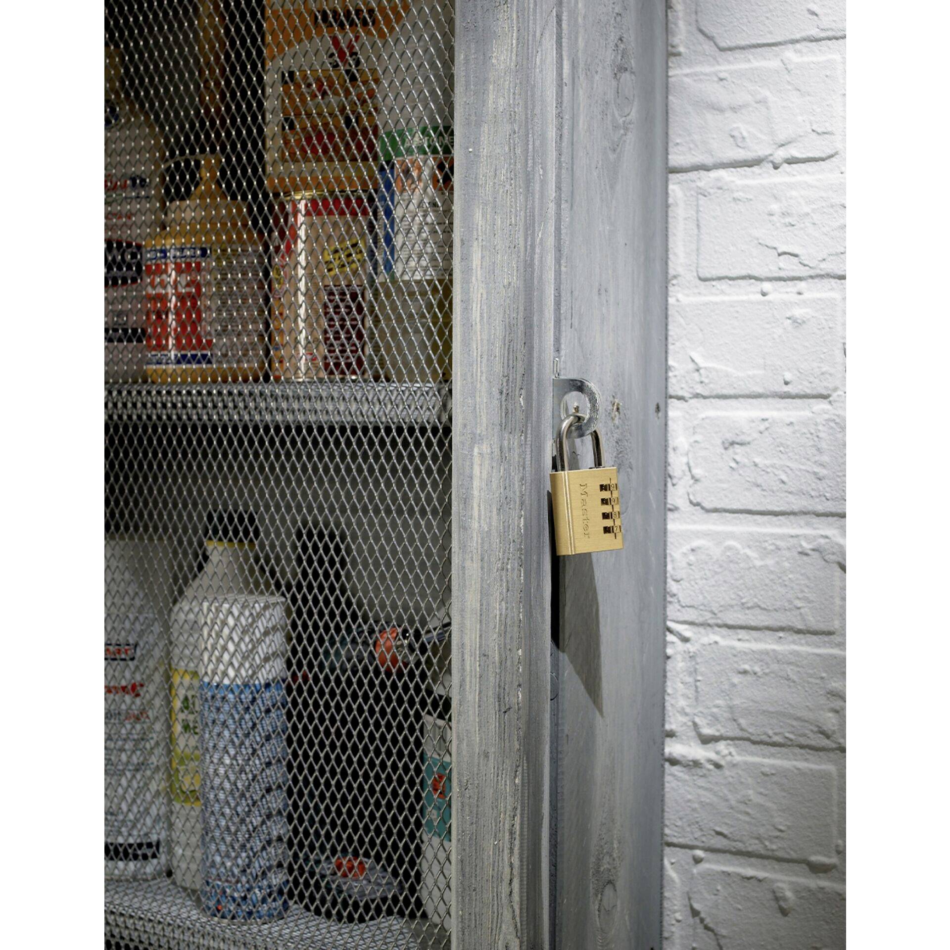 A metal cabinet with a mesh door stands against a white brick wall. The cabinet is filled with chemical bottles and secured with a combination lock.