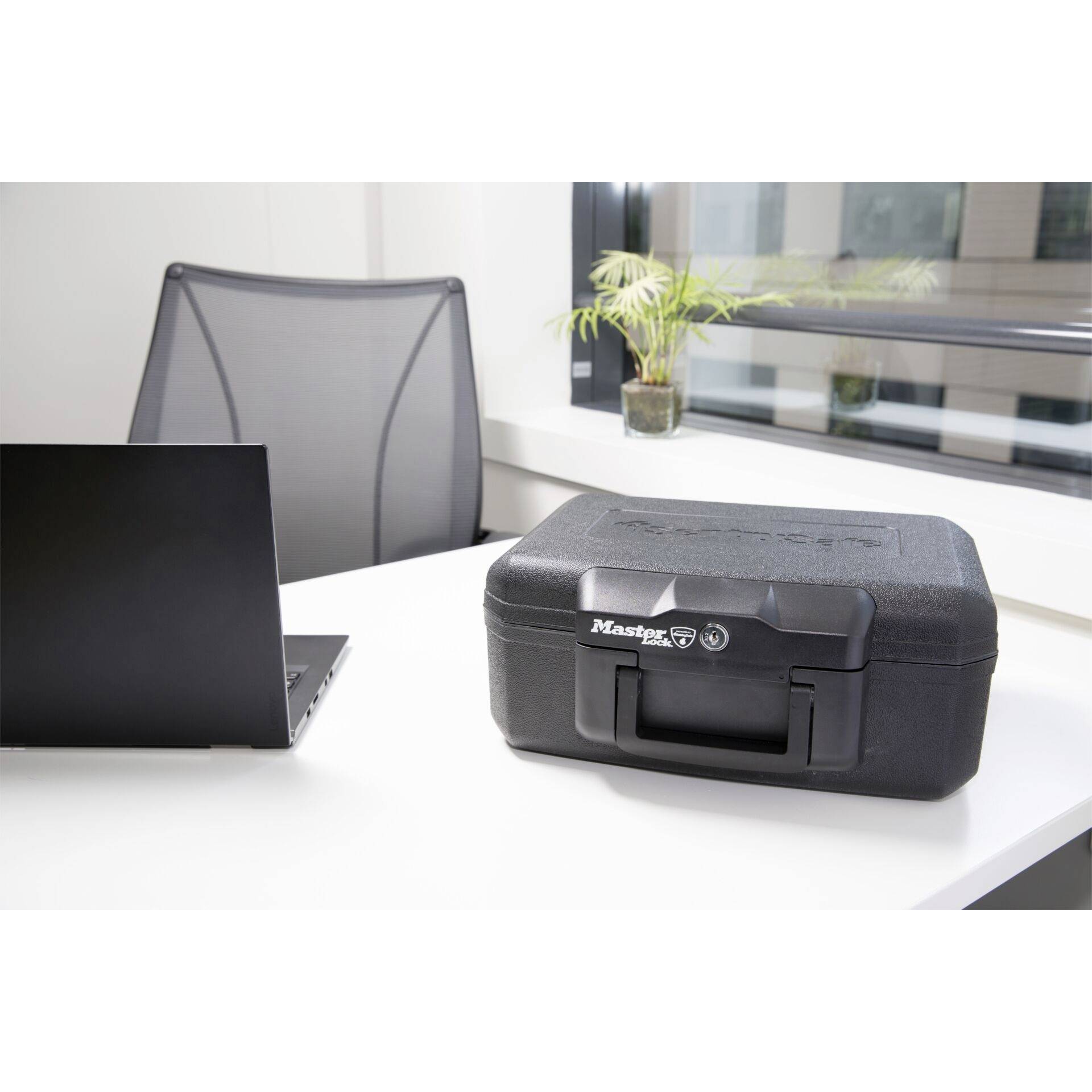 A black laptop sits on a white table next to a closed, black security case. In the background, there is an office chair and a window with plants.