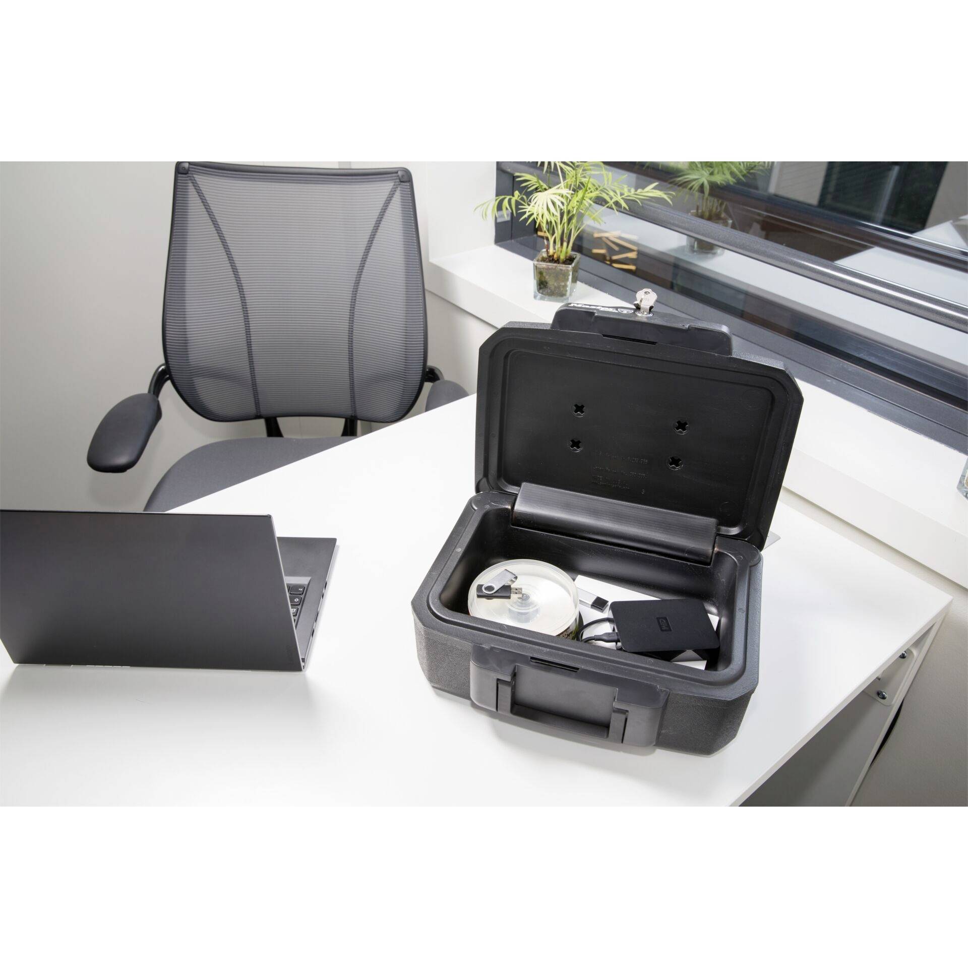 On a white desk sits an open black suitcase with technical equipment and a laptop. A chair is in the background.