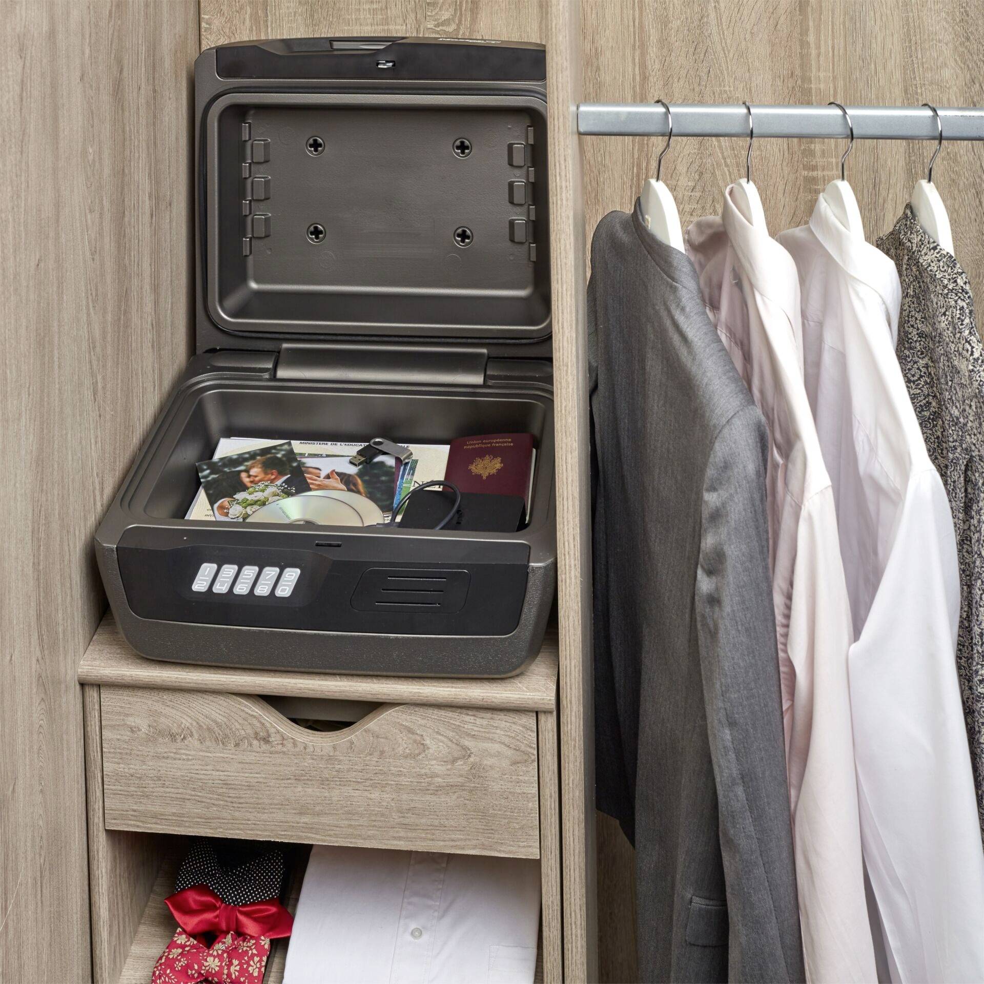 An open safe with documents, a passport and a CD is located on a chest of drawers next to several hanging shirts.
