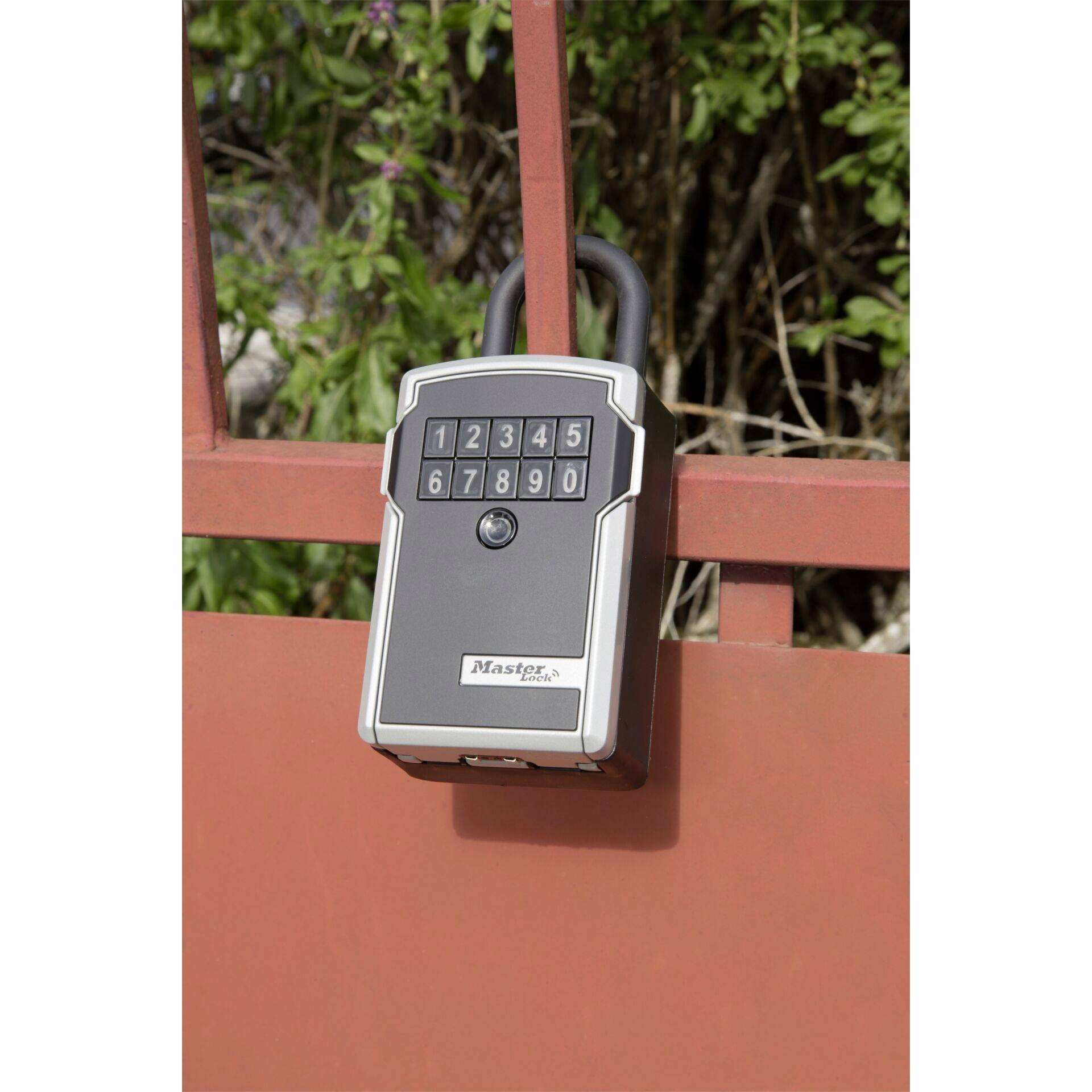 A combination lock is attached to a red metal gate. The lock has a numeric keypad ranging from 0 to 9. Plants are visible in the background.