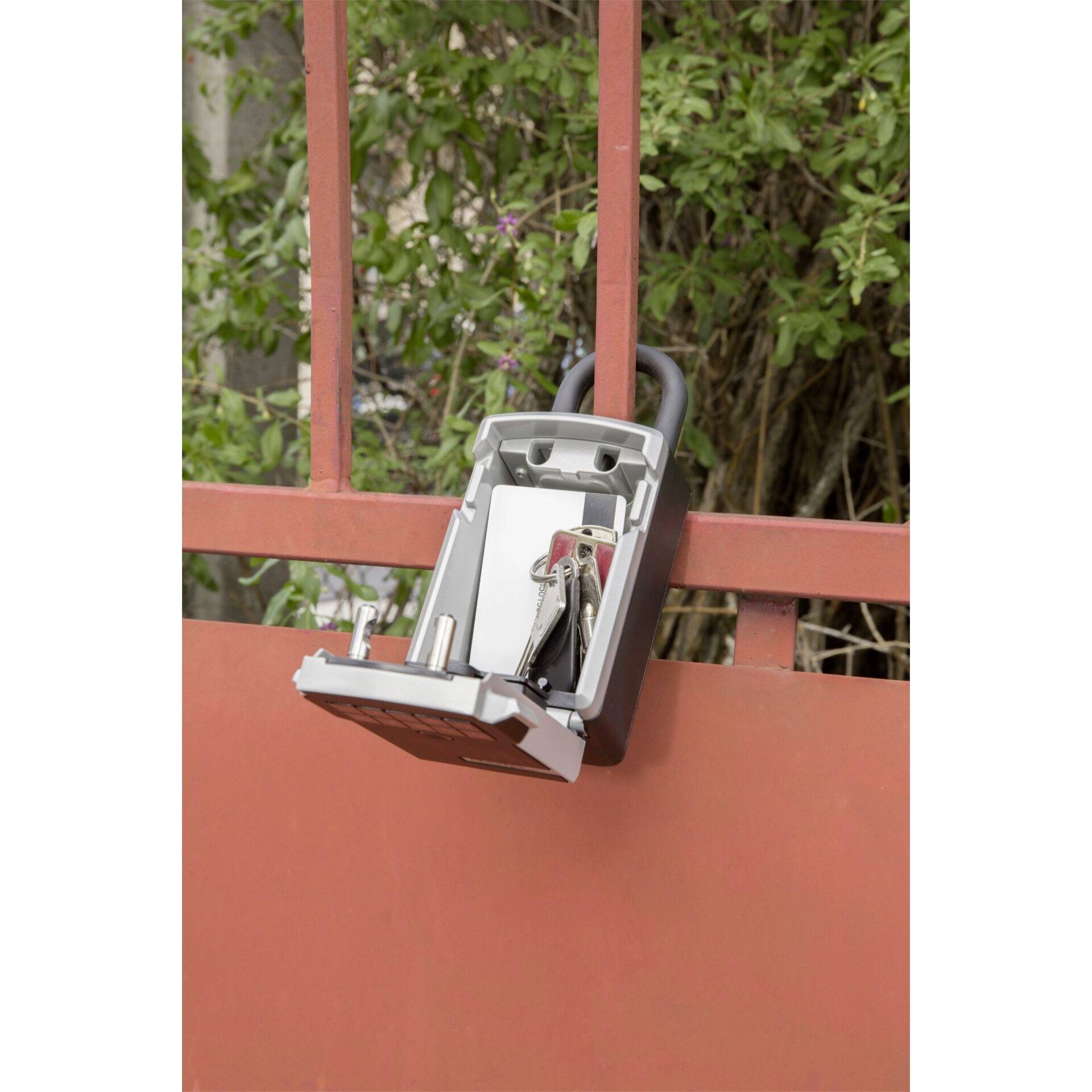 An open key box is hanging on a red metal fence. Several keys are hanging inside it. Green vegetation is visible in the background.