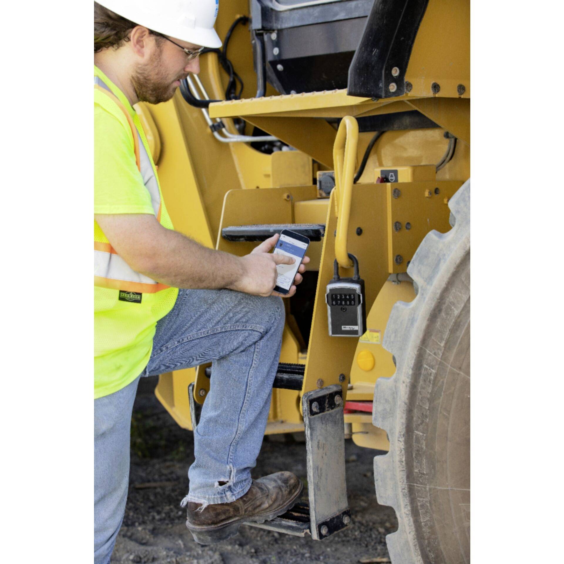 A construction worker in a high-visibility vest and hard hat stands next to a heavy-duty construction machine, checking something on a tablet.