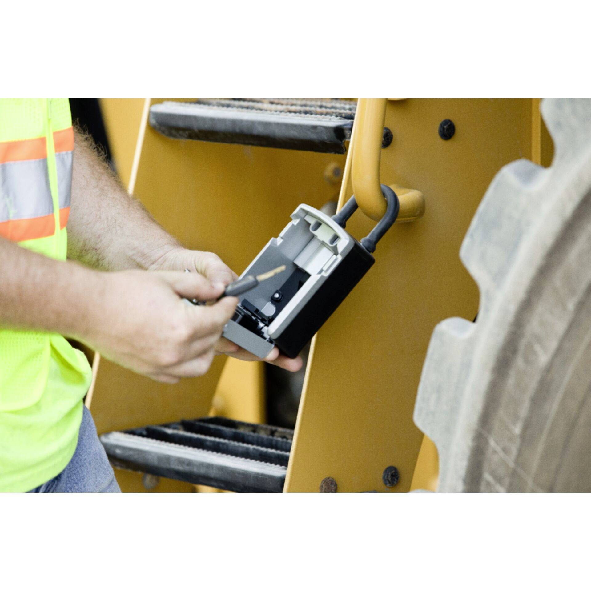 A person is inspecting an electrical device on a heavy vehicle, wearing a high-visibility vest. The focus is on safety inspection and maintenance.