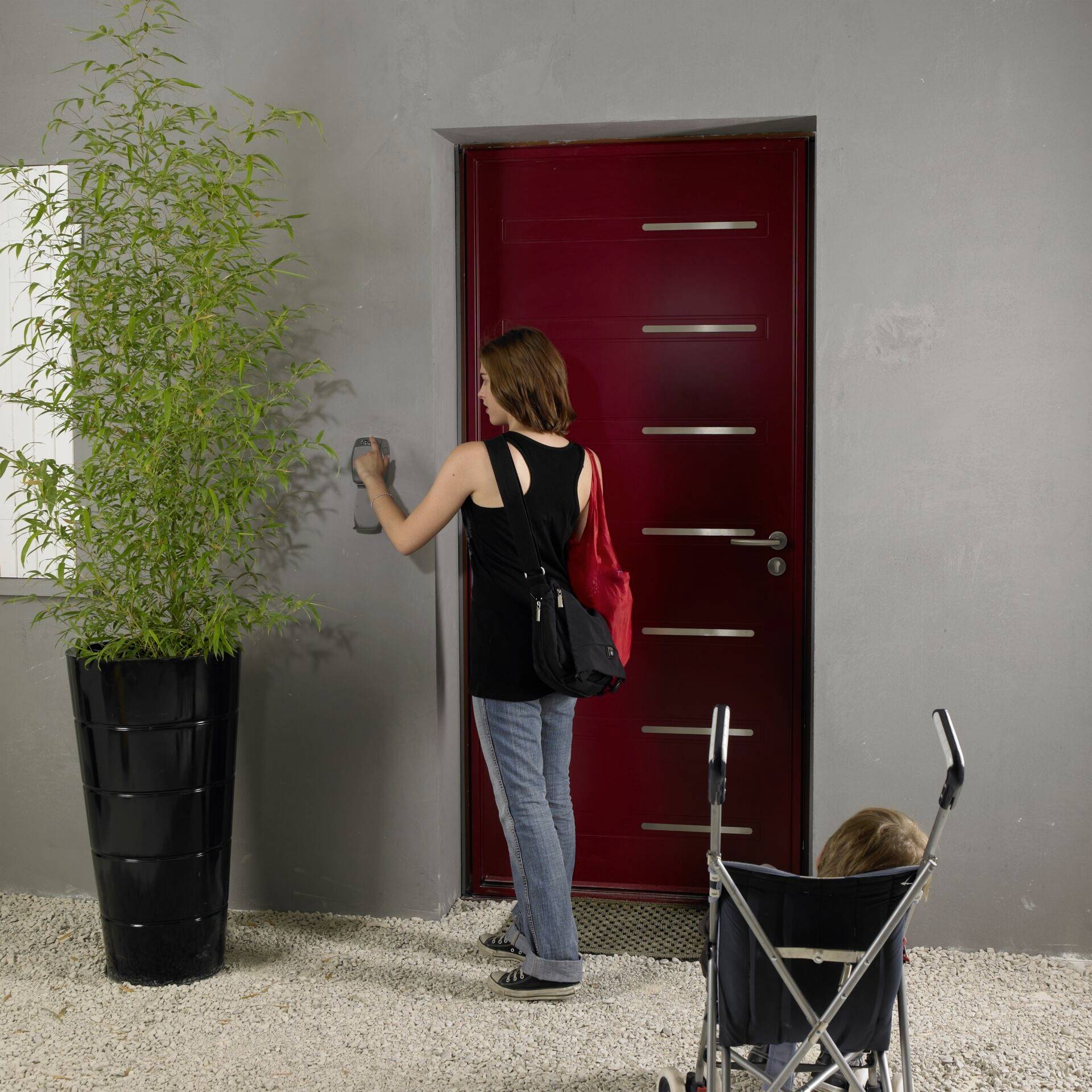 A woman is standing at a red door and ringing the doorbell. She is carrying a bag; next to her, a toddler sits in a pushchair. Plant on the left.