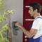 A man in workwear is repairing a grey wall switch. A plant is standing to the left, and a red wall is to the right.
