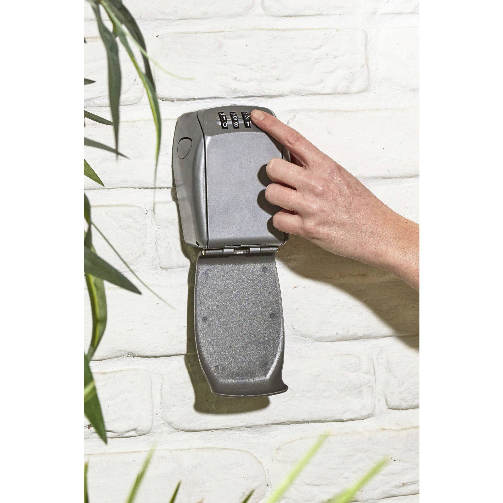A hand opens a key safe with a number combination, which is mounted on a white brick wall.