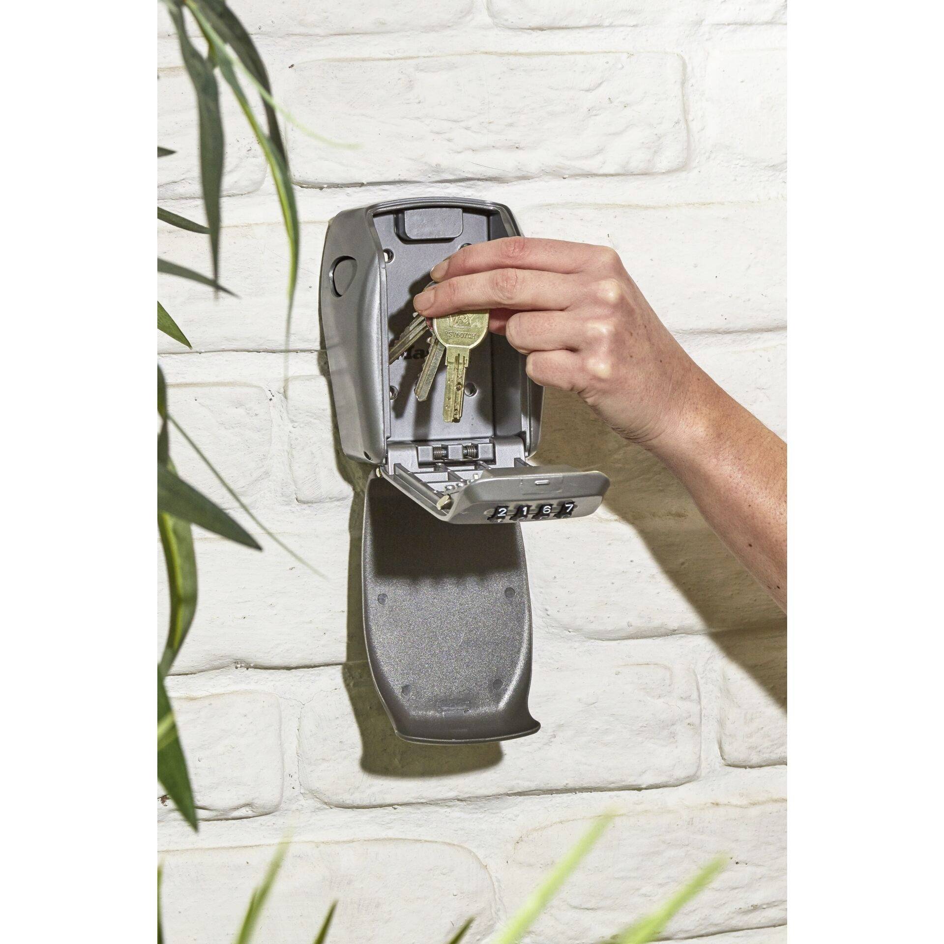 A hand is placing keys into a wall safe with a combination lock, which is mounted on a brick wall. Plant parts are visible on the left side.