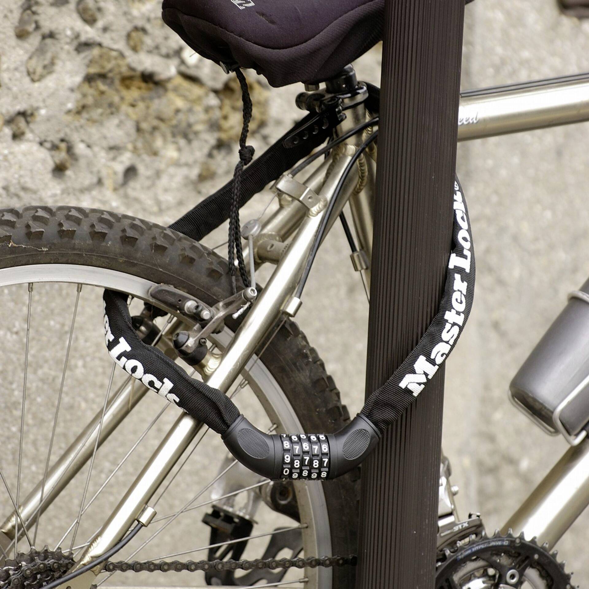 A silver bicycle is secured with a black combination lock to a metal post. A stone wall is visible in the background.