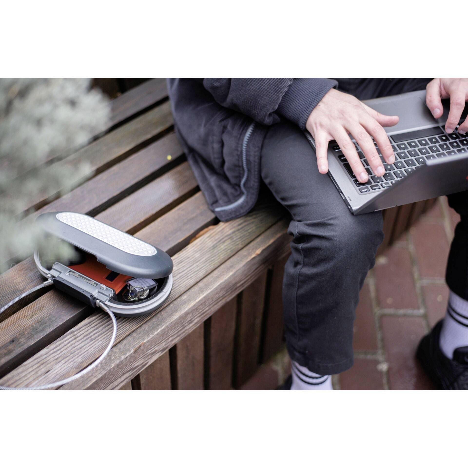 A person is sitting on a wooden bench and working on a laptop. Beside them lies an open hard drive enclosure with a visible hard drive.