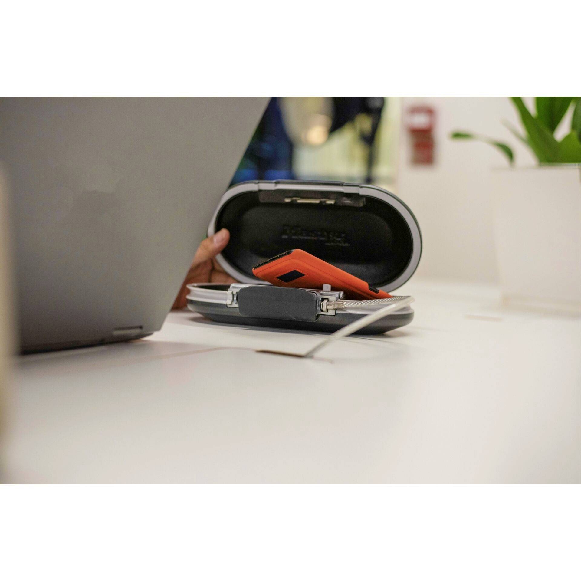 A person places an orange device into an oval box on a table. A plant stands beside it.