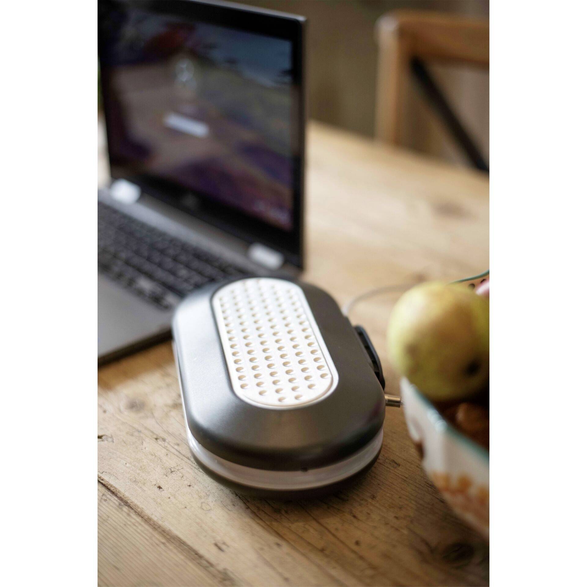 An electric bread bin sits on a wooden table next to an open laptop. In the background, a bowl of fruit can be seen.